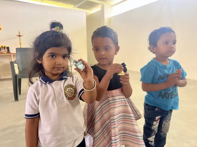 Two small girls in school uniform hold up little toys while a toddler watches, soft daylight