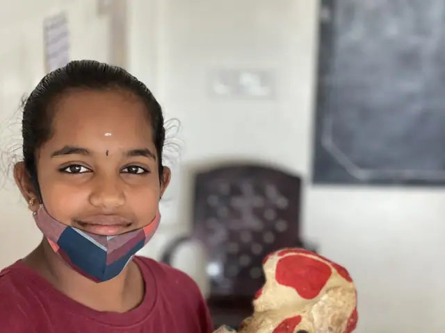 A girl in a maroon T-shirt smiles beside a coloured anatomical bone model on a stand