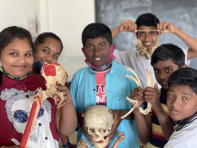 Six schoolchildren crowd together holding a human skull and anatomical bone models up to the camera