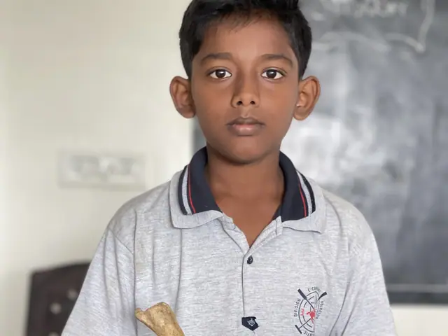 A boy at a wooden desk with a human skull and long bone laid out in front of him, chalkboard behind