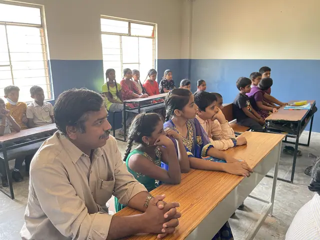 A teacher at a desk talks with two girls at the front of a bright classroom while the class works