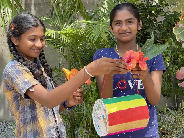 Two teenage girls in a garden hold a hand-painted drum decorated with hibiscus, smiling at the camera