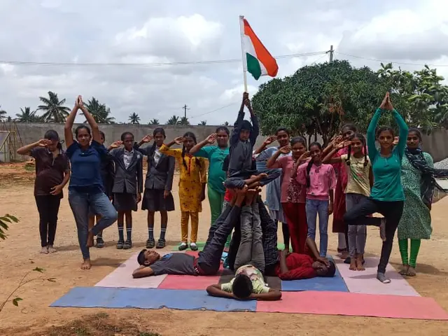 Yoga-day session on red earth: children holding poses on coloured mats with the Indian flag and a school bus behind