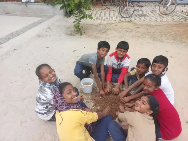 Ten children kneeling in a ring with their hands buried together in a mound of wet soil, several looking up grinning