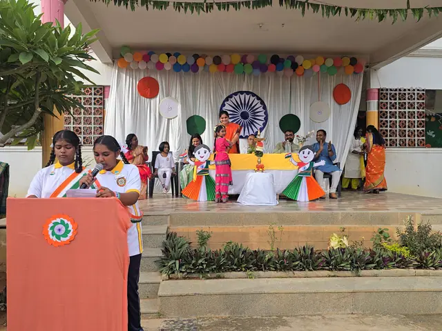 A schoolgirl speaks from a lectern draped in orange with a chakra emblem, students behind her