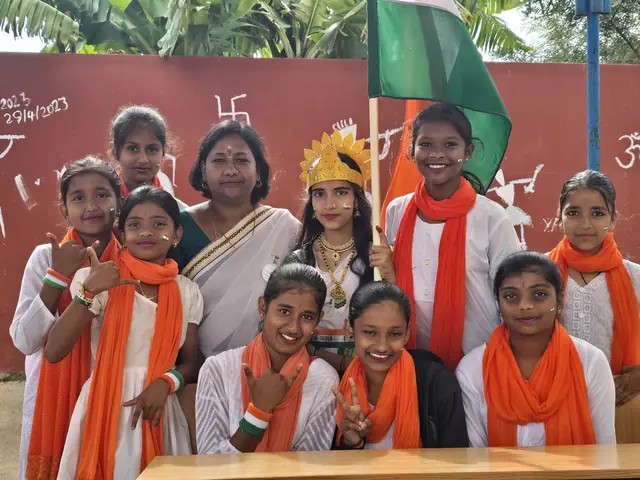 A teacher in a saree among five girls in orange-and-white costumes against a red mural wall