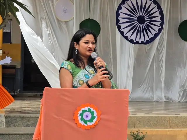 A woman in a green printed blouse speaks into a mic beside the orange lectern