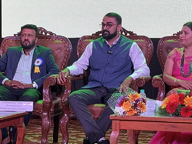 Three seated dignitaries in ornate chairs, warm stage lighting and floral foreground