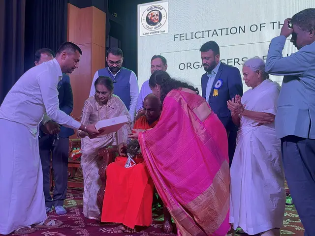 Guests present a shawl and a gift to an elderly honouree on stage, several hands reaching in