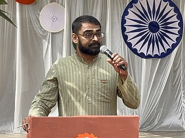 A man in a pale green kurta speaks into a handheld mic beside the chakra roundel