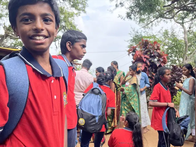 A boy in red uniform with a blue backpack grinning broadly at the camera, classmates resting behind