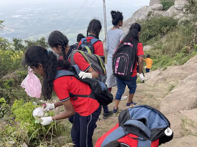 A girl crouches on a rocky path stuffing collected rubbish into a bag while others work up the slope