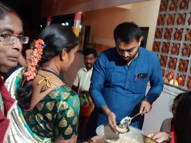 A woman in a garland in the foreground while a man serves food from a pot to a crowd in a hall