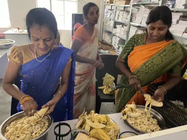 Two women in bright saris at a steel table peeling and slicing fruit into large trays