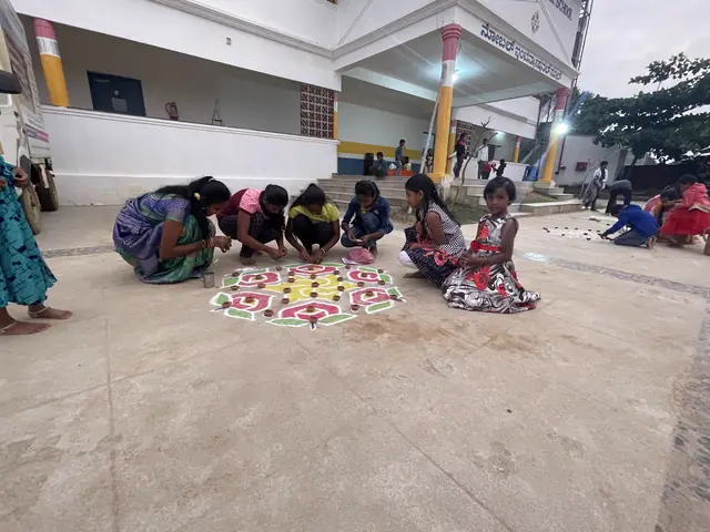 A woman and two girls kneel to fill in a bright rangoli with coloured powder outside the school portico