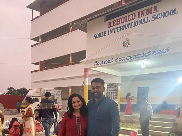 A couple in festive dress in front of the school block with the Rebuild India name board legible above