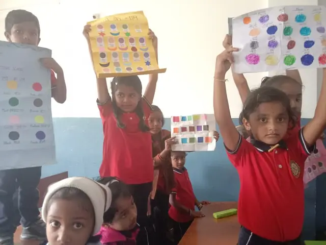 Children in red uniform hold hand-made colour charts up towards the camera, soft window light