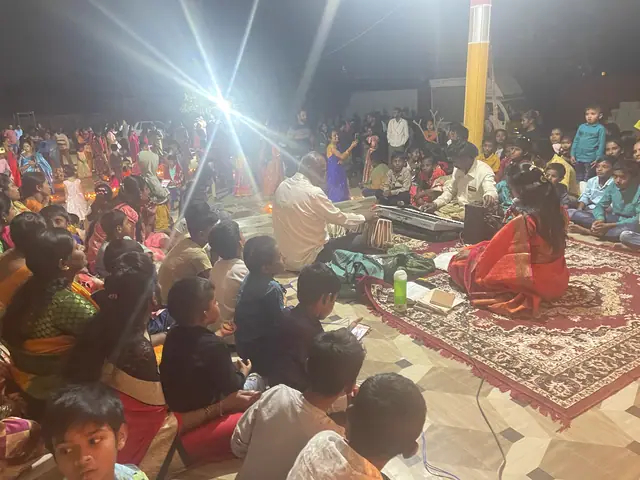 A large seated gathering of women in bright saris and children on carpets under a pillared hall at night