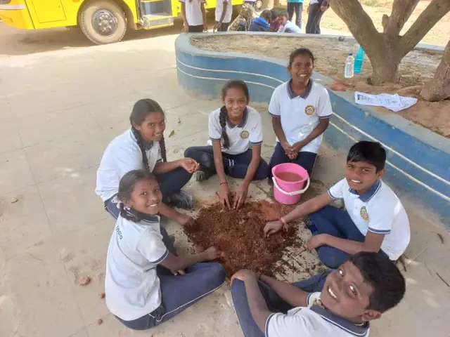 Four children kneeling around a heap of wet soil beside a concrete planter, school bus behind