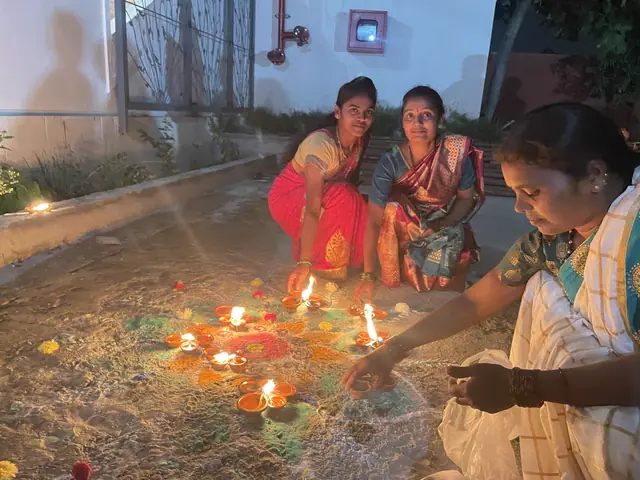 Four women in bright saris crouched together lighting a dense cluster of oil lamps at dusk