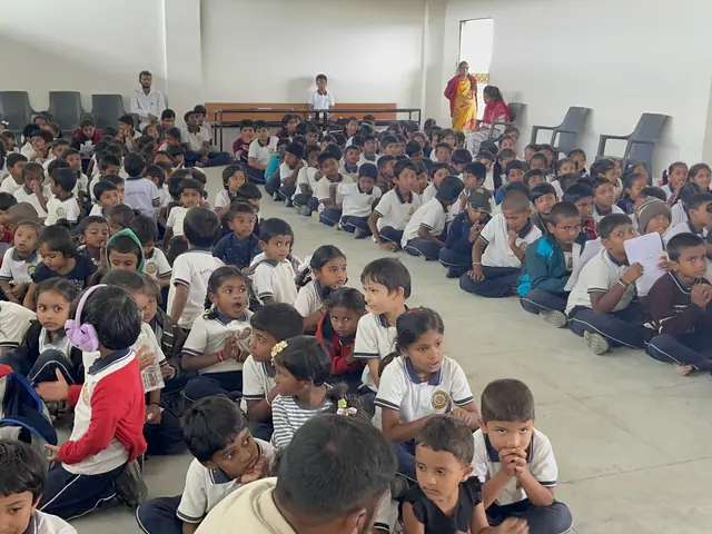 Tight rows of primary-age children sitting cross-legged on the hall floor, many faces turned to camera