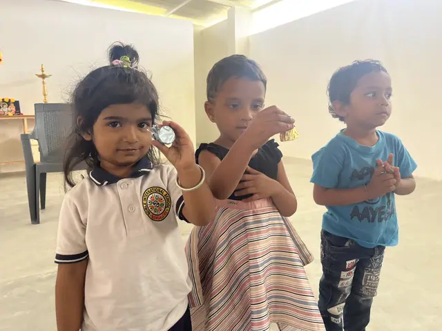 Three little children in a bright room, one holding up a small object towards the camera