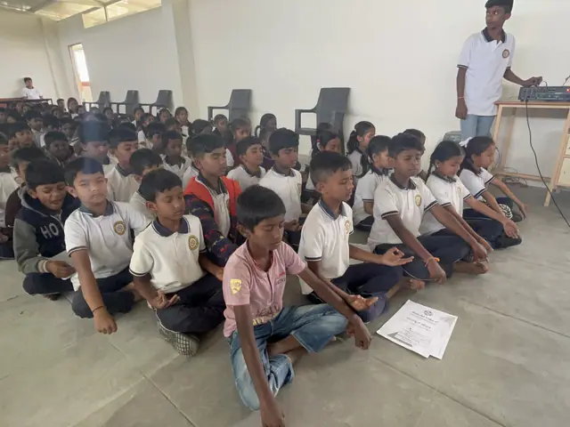 Rows of children seated on the floor with worksheets in front of them, a teacher at a table behind