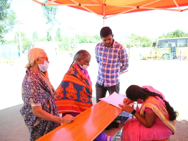 Two village women at an orange trestle registration desk under a red canopy, a volunteer writing