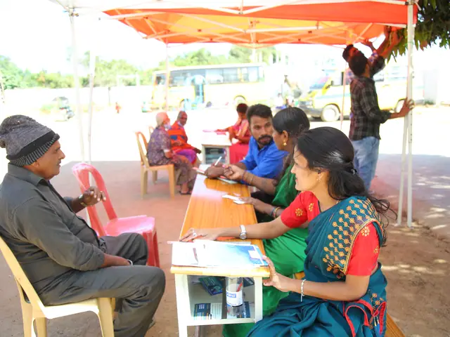 An elderly man seated at a health-camp desk with two women staff recording his details