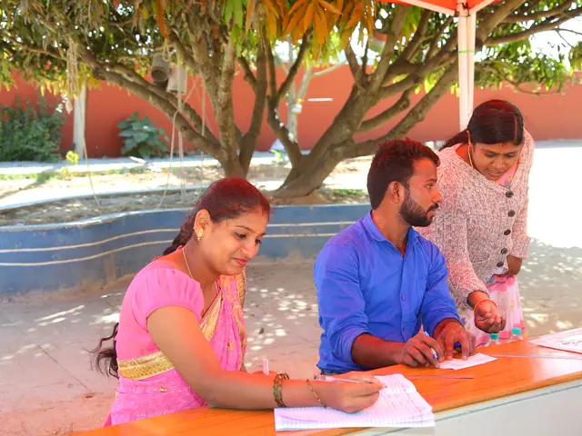 A health worker and two women in saris at a small table under a spreading tree, filling in forms
