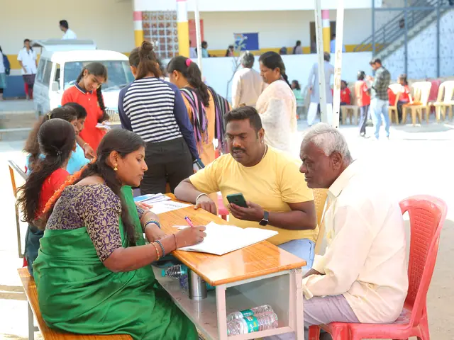 An older man seated across a desk from two women registering him, camp crowd blurred behind