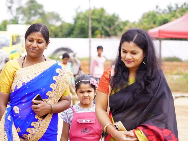 Two women in saris standing either side of a small girl in a pink pinafore at an outdoor event