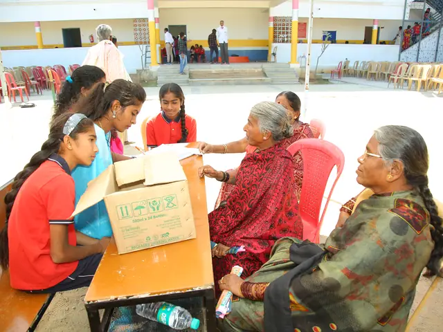 Two schoolgirls lean over a box to hand something to two elderly women seated outdoors