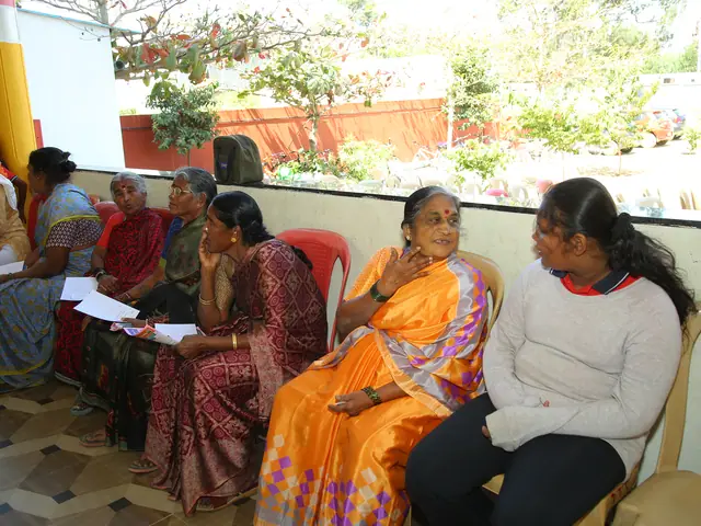 A row of village women sit waiting along a wall holding paper forms