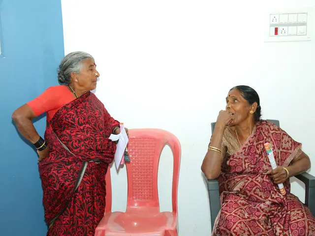 Two elderly women in deep red saris wait beside empty plastic chairs against a plain white wall