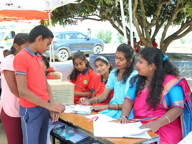 A boy carries a cardboard box past a table where girls fill in forms under a tree