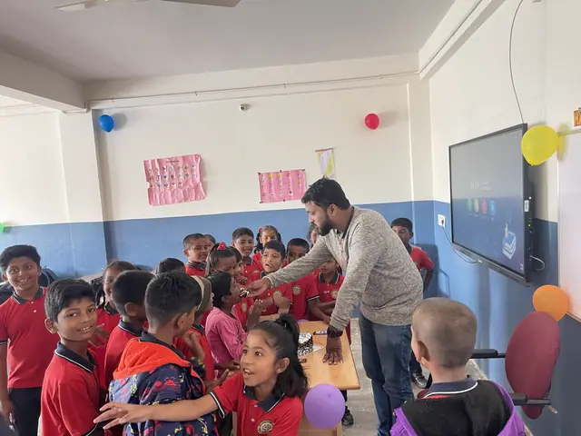 A teacher leans over small children in red uniform seated at desks beside a wall-mounted smartboard