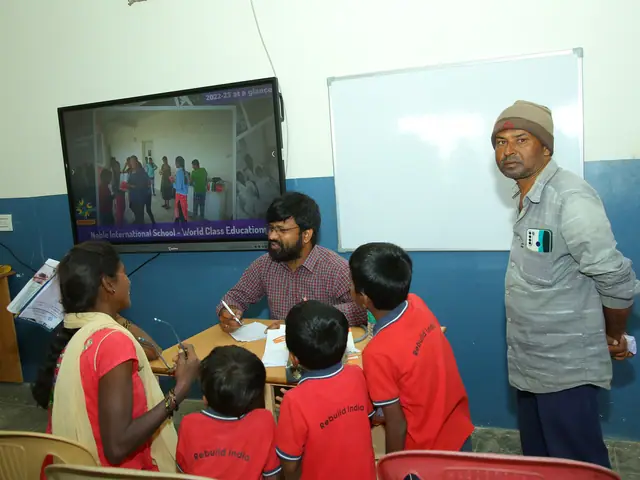 Children in red uniform at a desk facing a wall-mounted screen, two adults at either side