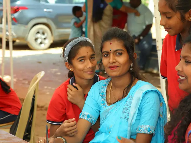 A teenage girl and a young woman sit close together outdoors and smile straight at the camera