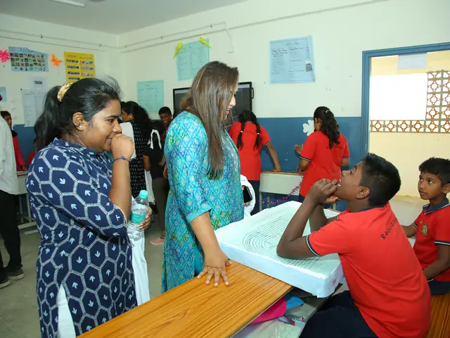 Children lean over a long washbasin trough in a colourful classroom while two women talk beside them