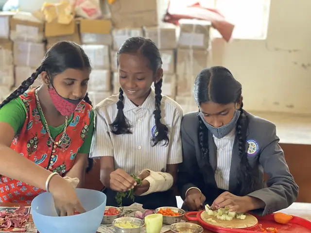 Three girls stand chopping fruit and vegetables at a table, all concentrating on their hands