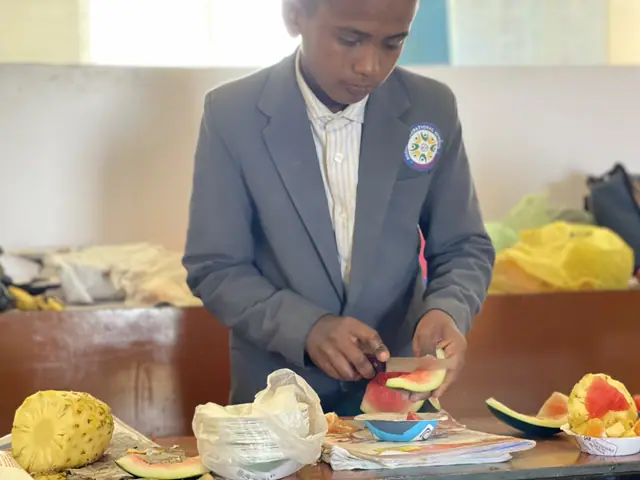 A boy in a school blazer works intently on a carved fruit arrangement laid out on a table
