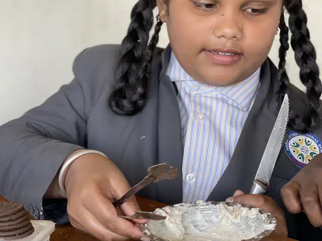 A girl with bows in her hair lifts a spoon over a plate and glances up towards the camera