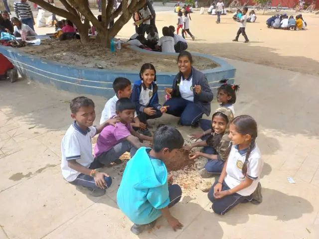 Children in white uniforms sitting cross-legged on sandy ground around a circular blue tree planter