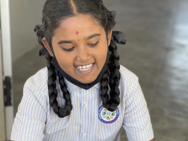 A girl with beribboned plaits sits peeling a cucumber and breaks into a wide grin