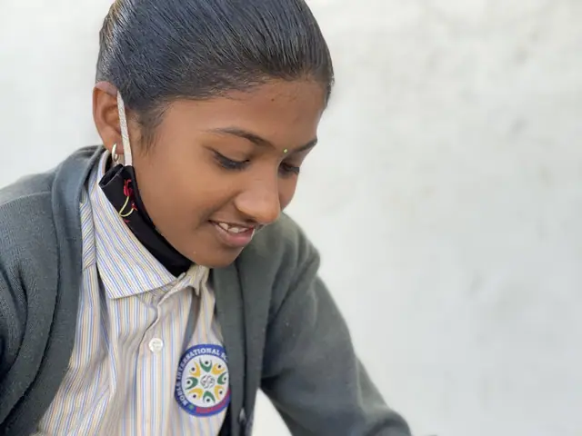 A smiling girl in a grey school sweater sits cross-legged on a verandah peeling fruit