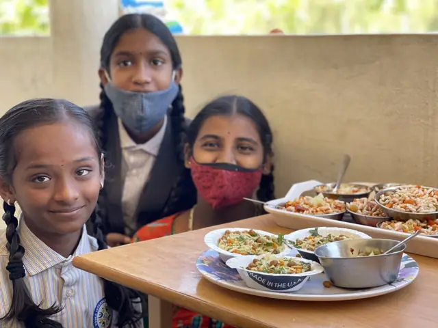 Three schoolgirls behind a table laid with plates of food lean in together and smile at the camera