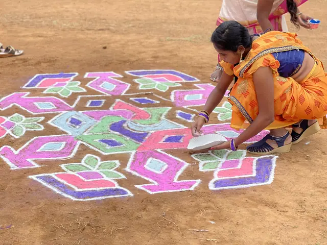 A woman in an orange and pink sari crouches on bare earth filling in a huge geometric rangoli