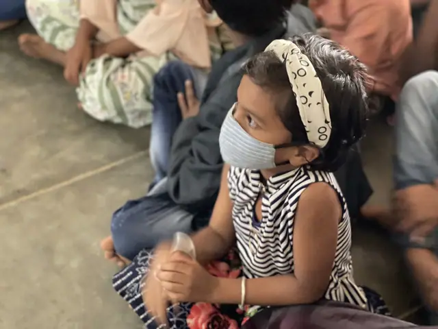 A girl sits cross-legged among other children, hands folded, watching something off frame