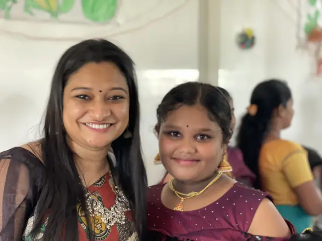 A young woman and a teenage girl lean together and smile straight at the camera